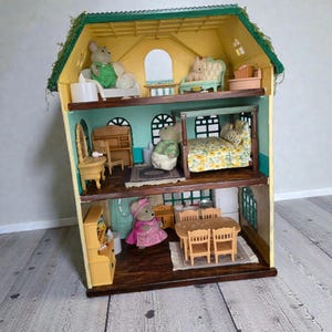 K&ouml;nnte beinhalten: Ein mehrst&ouml;ckiges Puppenhaus mit gelber Au&szlig;enfassade und gr&uuml;nem Dach, eingerichtet mit Miniaturm&ouml;beln und Figuren. Das Puppenhaus verf&uuml;gt &uuml;ber mehrere Zimmer, darunter ein Schlafzimmer, ein Wohnzimmer und ein Essbereich, die jeweils mit winzigen Accessoires dekoriert sind. Ein bezauberndes Spielzeug.