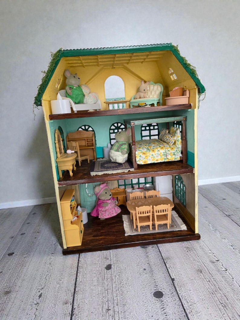 K&ouml;nnte beinhalten: Ein mehrst&ouml;ckiges Puppenhaus mit gelber Au&szlig;enfassade und gr&uuml;nem Dach, eingerichtet mit Miniaturm&ouml;beln und Figuren. Das Puppenhaus verf&uuml;gt &uuml;ber mehrere Zimmer, darunter ein Schlafzimmer, ein Wohnzimmer und ein Essbereich, die jeweils mit winzigen Accessoires dekoriert sind. Ein bezauberndes Spielzeug.
