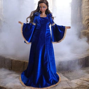 Può includere: Una bambola vestita con un abito di velluto blu con bordi dorati e una lunga parrucca castano fluente. La bambola è in piedi su una scalinata di pietra in un'ambientazione nebbiosa ed eterea.