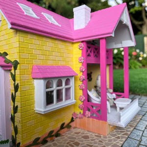 May include: A miniature dollhouse with a yellow brick exterior, a pink roof, and a white chimney. The house features a porch with a white railing and pink columns, and is decorated with pink roses. A small window is visible.