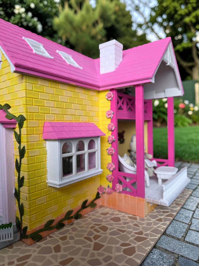 May include: A miniature dollhouse with a yellow brick exterior, a pink roof, and a white chimney. The house features a porch with a white railing and pink columns, and is decorated with pink roses. A small window is visible.