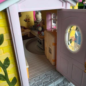 May include: A miniature dollhouse with a yellow brick exterior, pink trim, and a partially open door. The interior reveals a glimpse of furnished rooms, including a table and a suitcase. The door has a small, oval window.