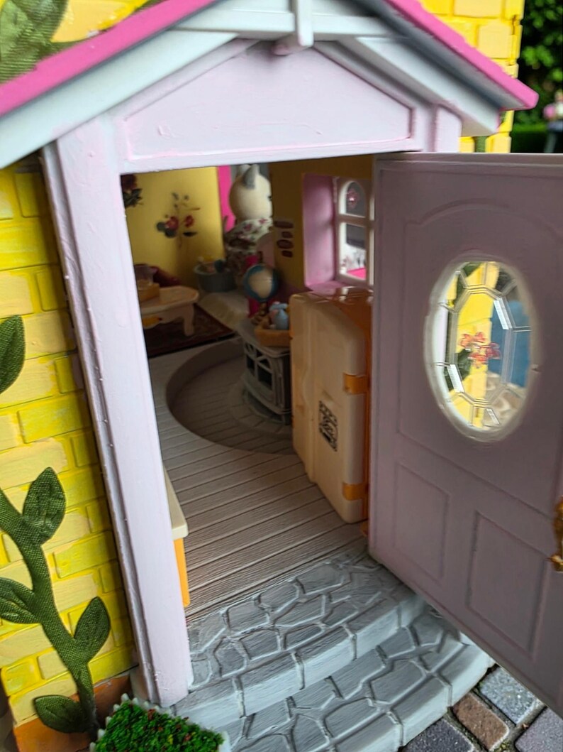 May include: A miniature dollhouse with a yellow brick exterior, pink trim, and a partially open door. The interior reveals a glimpse of furnished rooms, including a table and a suitcase. The door has a small, oval window.