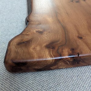 K&ouml;nnte beinhalten: Nahaufnahme eines Holzservierbretts mit nat&uuml;rlicher Kante. Das Brett ist tiefbraun mit sichtbarer Holzmaserung und Knoten. Die Oberfl&auml;che ist glatt und poliert und reflektiert das Licht. Das Brett wird wahrscheinlich zum Servieren von Speisen verwendet.