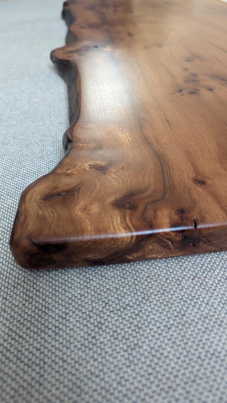 K&ouml;nnte beinhalten: Nahaufnahme eines Holzservierbretts mit nat&uuml;rlicher Kante. Das Brett ist tiefbraun mit sichtbarer Holzmaserung und Knoten. Die Oberfl&auml;che ist glatt und poliert und reflektiert das Licht. Das Brett wird wahrscheinlich zum Servieren von Speisen verwendet.