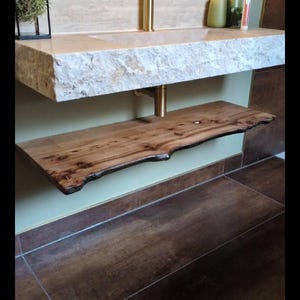 K&ouml;nnte beinhalten: Ein modernes Badezimmer-Waschbecken mit einer Steinplatte und einem Regal aus Holz mit lebendiger Kante. Der Wasserhahn und die Halterung sind goldfarben. Die W&auml;nde sind hellgr&uuml;n gestrichen und der Boden ist mit braunen Fliesen gefliest.