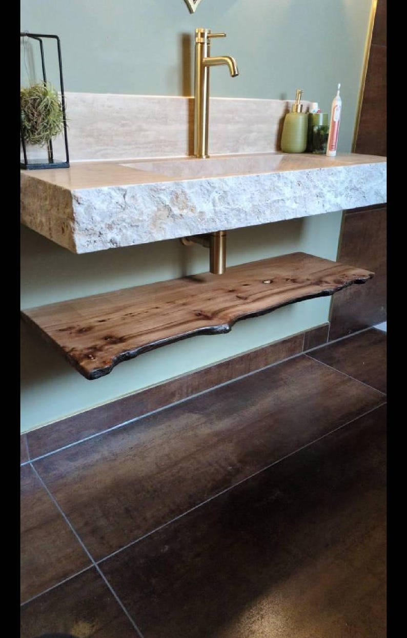 K&ouml;nnte beinhalten: Ein modernes Badezimmer-Waschbecken mit einer Steinplatte und einem Regal aus Holz mit lebendiger Kante. Der Wasserhahn und die Halterung sind goldfarben. Die W&auml;nde sind hellgr&uuml;n gestrichen und der Boden ist mit braunen Fliesen gefliest.