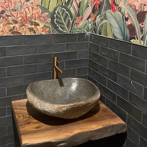 Könnte beinhalten: Ein Steinwaschbecken mit einer bronzefarbenen Armatur steht auf einem Waschtisch aus Holz mit lebendiger Kante. Das Waschbecken ist grau und hat eine natürliche, organische Form. Die Wand ist mit dunkelgrauen rechteckigen Fliesen gefliest, und die Tapete hat ein farbenfrohes Blumenmuster.