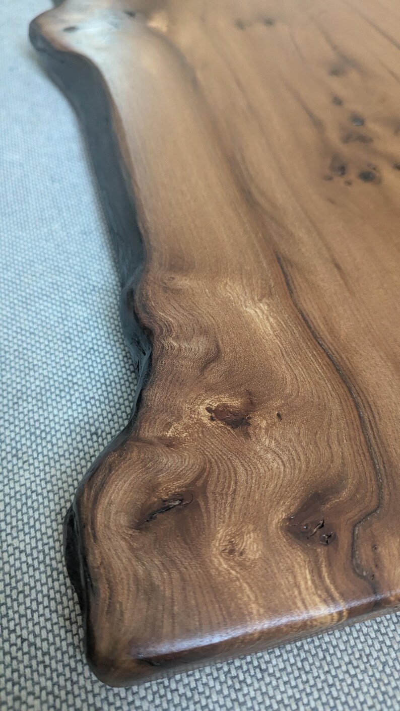 K&ouml;nnte beinhalten: Ein Holzservierbrett mit nat&uuml;rlicher Kante. Das Brett hat eine warme braune Farbe mit dunkleren Knoten und Maserungsmustern. Der Rand ist dunkelbraun und die Oberfl&auml;che ist glatt und poliert. Ideal zum Servieren von Vorspeisen oder Charcuterie.