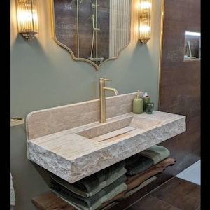K&ouml;nnte beinhalten: Ein modernes Badezimmer-Waschbecken mit einem Steinwaschbecken und einer goldenen Armatur. Das Waschbecken hat eine nat&uuml;rliche Steinoptik und ist auf einem Holzregal mit gefalteten Handt&uuml;chern montiert. Zwei Wandleuchten flankieren einen dekorativen Spiegel.