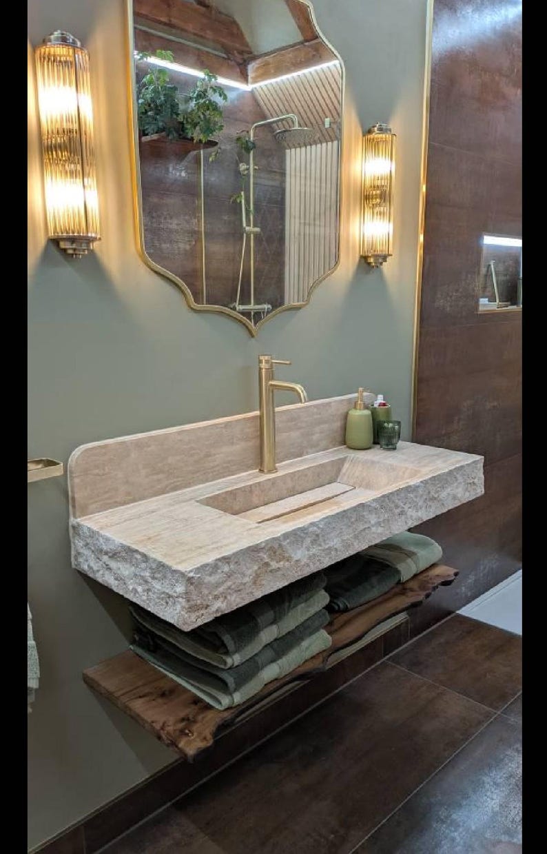 K&ouml;nnte beinhalten: Ein modernes Badezimmer-Waschbecken mit einem Steinwaschbecken und einer goldenen Armatur. Das Waschbecken hat eine nat&uuml;rliche Steinoptik und ist auf einem Holzregal mit gefalteten Handt&uuml;chern montiert. Zwei Wandleuchten flankieren einen dekorativen Spiegel.
