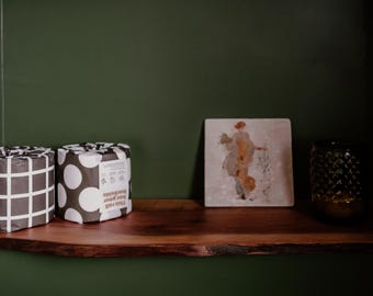 May include: A wooden shelf displays two rolls of toilet paper with black and white patterned wrapping, a framed artwork of a person, and a decorative glass jar. The shelf is against a dark green wall.