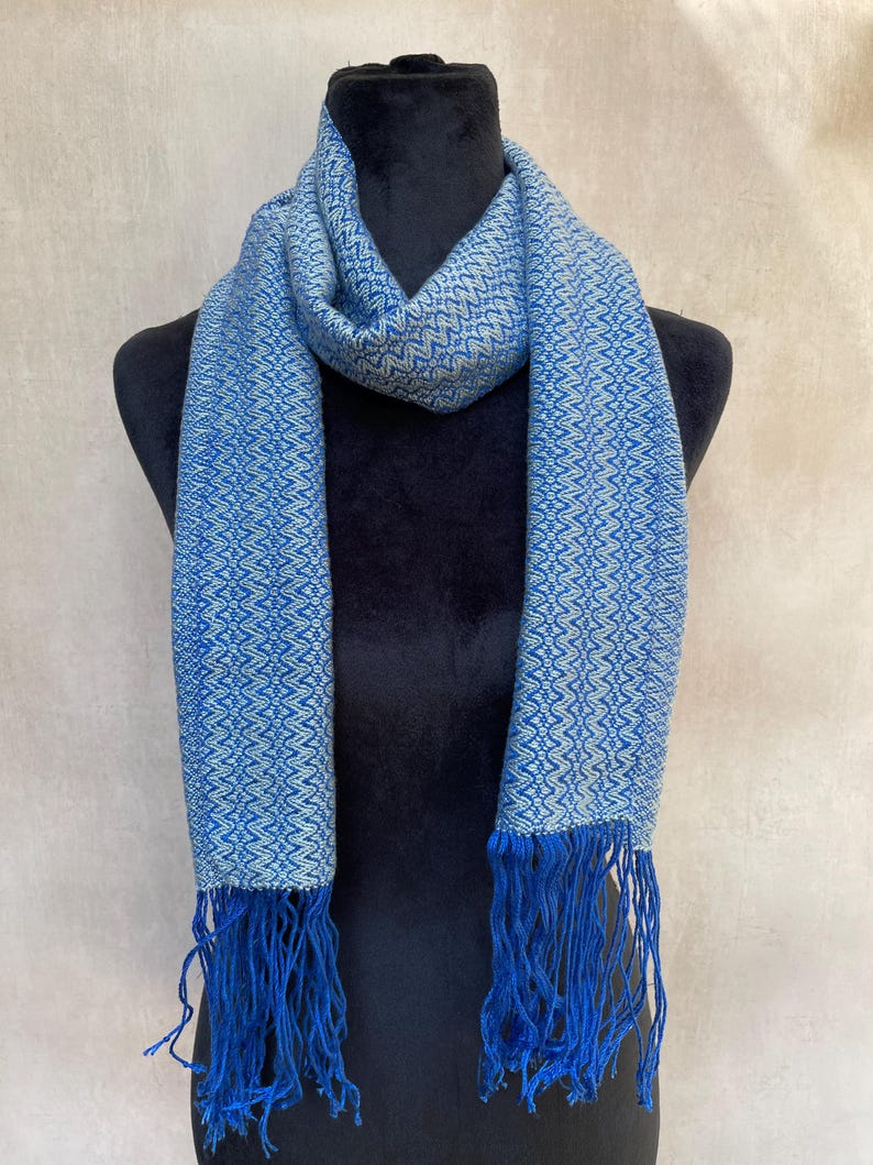 May include: A blue scarf with a geometric pattern and fringe. The scarf is displayed on a black mannequin. The scarf's pattern is a lighter blue, creating a contrast with the darker blue background.