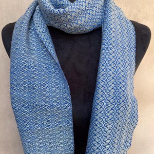 May include: A blue and white patterned scarf with fringe. The scarf is displayed on a black mannequin. The scarf has a woven design with a repeating geometric pattern. The fringe is a darker shade of blue.