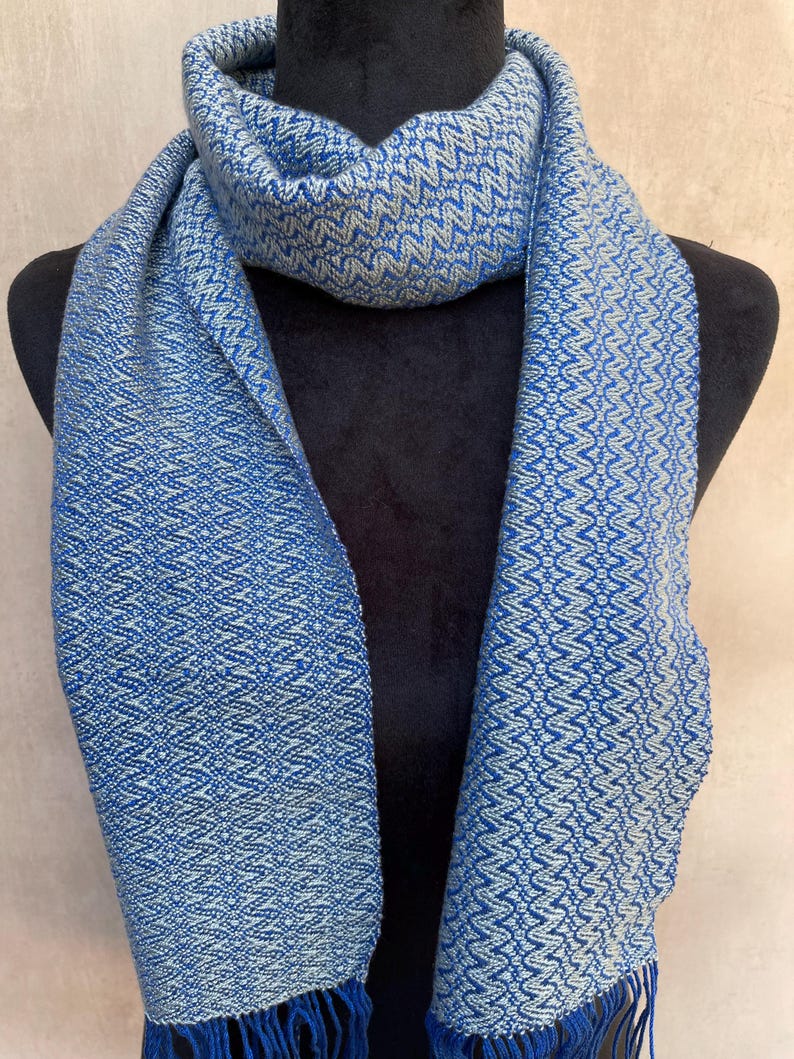 May include: A blue and white patterned scarf with fringe. The scarf is displayed on a black mannequin. The scarf has a woven design with a repeating geometric pattern. The fringe is a darker shade of blue.