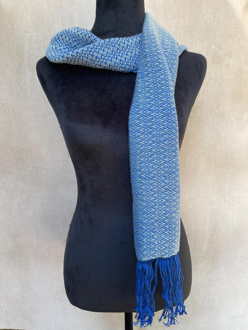 May include: A blue and white patterned scarf with fringe. The scarf is draped over a black mannequin. The scarf has a geometric design and is made of a soft material. The fringe is a darker shade of blue.
