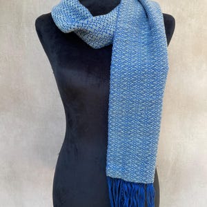 May include: A blue and white woven scarf with a geometric pattern and fringe. The scarf is draped over a black mannequin. The scarf's color is a mix of light and dark blue, creating a textured appearance. The fringe is a darker shade of blue.