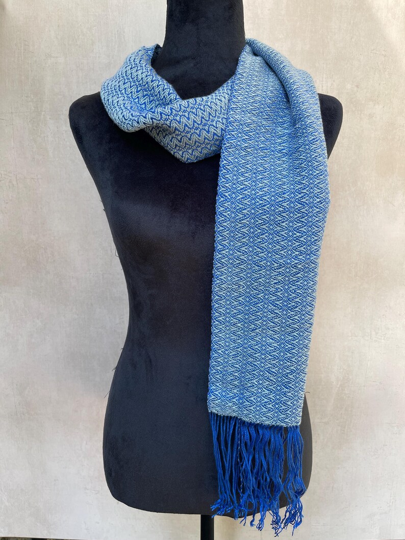 May include: A blue and white woven scarf with a geometric pattern and fringe. The scarf is draped over a black mannequin. The scarf's color is a mix of light and dark blue, creating a textured appearance. The fringe is a darker shade of blue.
