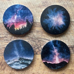 Puede incluir: Cuatro posavasos redondos con diseños celestiales. Cada posavasos presenta una pintura de acuarela diferente de un cielo nocturno con estrellas, montañas y árboles. La paleta de colores incluye azules, morados y rosas.