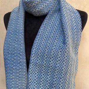 May include: A blue and white patterned scarf with a fringed edge. The scarf is displayed on a black mannequin. The scarf has a woven design with a repeating zig-zag pattern. The color is a medium blue with white accents.