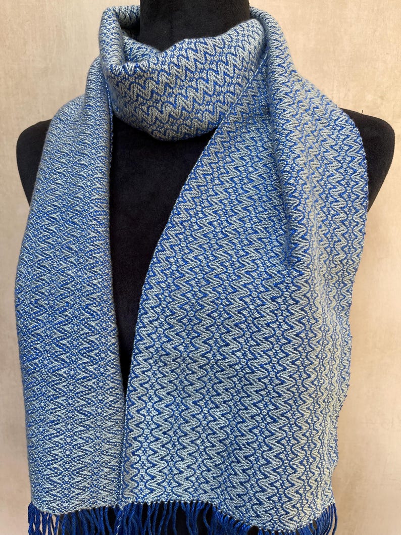 May include: A blue and white patterned scarf with a fringed edge. The scarf is displayed on a black mannequin. The scarf has a woven design with a repeating zig-zag pattern. The color is a medium blue with white accents.