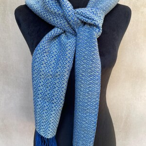 May include: A blue and white patterned scarf with fringe. The scarf is displayed on a black mannequin. The scarf has a woven design with a blue and white color scheme. The fringe is a darker shade of blue.