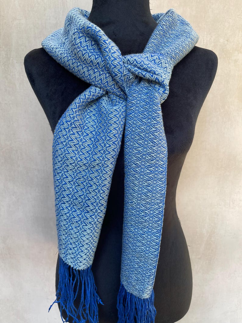 May include: A blue and white patterned scarf with fringe. The scarf is displayed on a black mannequin. The scarf has a woven design with a blue and white color scheme. The fringe is a darker shade of blue.