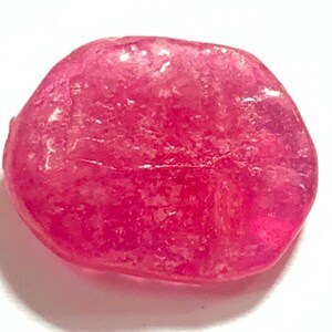 Könnte beinhalten: Ein polierter, oval geformter, transluzenter Edelstein in einem leuchtenden Rosaton. Der Stein hat eine glatte Oberfläche mit subtilen inneren Texturen, vor einem hellen weißen Hintergrund.