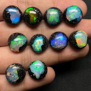 Può includere: Una collezione di undici pietre preziose di opale rotonde. Ogni pietra ha una base nera macchiettata con colori iridescenti vibranti, tra cui blu, verde e giallo. Gli opali sono presentati su uno sfondo scuro.