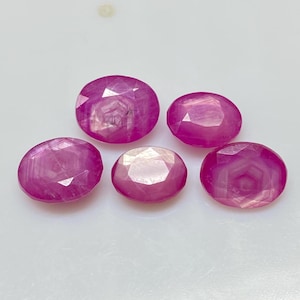 Könnte beinhalten: Fünf oval geschliffene, facettierte Edelsteine in verschiedenen Rosa- und Rottönen. Die Edelsteine sind auf einer weißen Oberfläche angeordnet und zeigen ihre polierten Oberflächen und inneren Strukturen. Bei den Edelsteinen handelt es sich wahrscheinlich um Rubine.