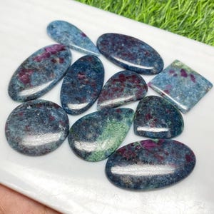 Könnte beinhalten: Eine Sammlung polierter Edelsteine in verschiedenen Formen, darunter oval, tropfenförmig und quadratisch. Die Steine weisen eine Mischung aus blau-grünen und roten Farbtönen mit glänzendem Finish auf. Sie sind auf einer weißen Oberfläche angeordnet.