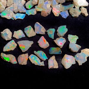 Może przedstawiać: Kolekcja surowych kamieni szlachetnych opalu o żywych, iryzujących kolorach. Opale wykazują błyski niebieskiego, zielonego, pomarańczowego i różowego w swojej naturalnej matrycy. Kamienie szlachetne są na czarnej powierzchni.