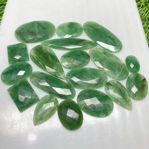 Könnte beinhalten: Eine Sammlung facettierter grüner Edelsteine in verschiedenen Formen, darunter Ovale, Rechtecke und Tropfen. Die Edelsteine sind auf einer weißen Oberfläche angeordnet, wodurch ihre polierten Oberflächen und inneren Muster hervorgehoben werden. Die Edelsteine variieren in der Größe und sind durchscheinend.