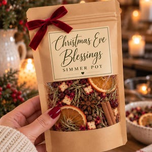 May include: A brown paper pouch with a clear window containing dried orange slices, cinnamon sticks, star anise, cranberries, and rosemary. A dark red velvet bow is attached to the top. The label reads Christmas Eve Blessings SIMMER POT.