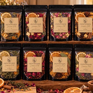 May include: Eight black pouches containing dried botanical blends labeled Intention Simmer Pot. Each pouch features a clear window displaying dried orange slices, cinnamon sticks, flower petals, and herbs with unique text labels for various intentions.
