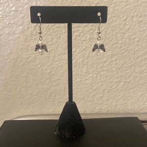 Puede incluir: Pendientes de plata con forma de alas de &aacute;ngel con una perla de cristal transparente y una perla blanca. Los pendientes est&aacute;n colgados de un soporte de pendientes negro.