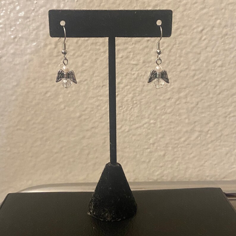 Puede incluir: Pendientes de plata con forma de alas de &aacute;ngel con una perla de cristal transparente y una perla blanca. Los pendientes est&aacute;n colgados de un soporte de pendientes negro.