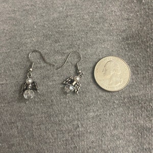 Puede incluir: Un par de pendientes de &aacute;ngel colgantes de plata con detalles de cristal transparente y una perla blanca en cada pendiente. Los pendientes se muestran junto a una moneda de veinticinco centavos de d&oacute;lar estadounidense para referencia de tama&ntilde;o.