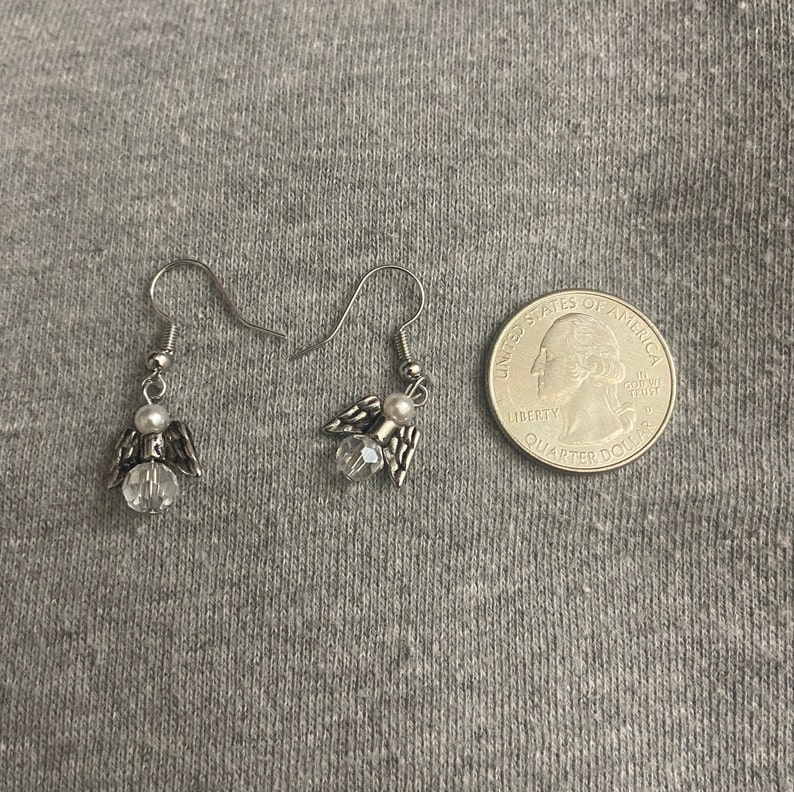 Puede incluir: Un par de pendientes de &aacute;ngel colgantes de plata con detalles de cristal transparente y una perla blanca en cada pendiente. Los pendientes se muestran junto a una moneda de veinticinco centavos de d&oacute;lar estadounidense para referencia de tama&ntilde;o.