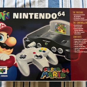 May include: Nintendo 64 game console box featuring Mario. The box shows the console, a controller, and a Super Mario 64 game cartridge. The box has the Nintendo 64 logo and text describing the console's features.
