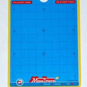 Puede incluir: Un tablero de juego azul y amarillo con un patrón de cuadrícula y el texto "Mine Storm" en rojo. El tablero tiene etiquetas para "Jugador Uno" y "Jugador Dos" e incluye las palabras "Escape", "Empuje" y "Fuego".