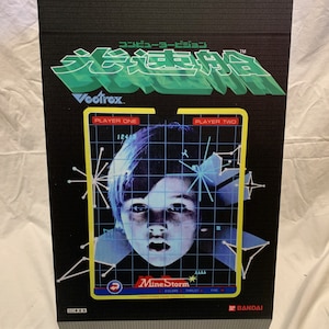 May include: A black and green box with a blue and white image of a child's face on the front. The box has the text "コンピュータービジョン 光速胜 Vectrex" in Japanese and the text "Mine Storm" in English. The box also has the text "BANDAI" in red.