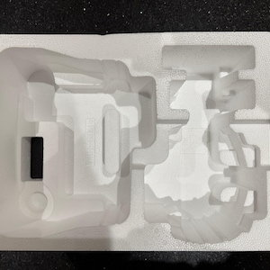 Könnte beinhalten: Weiße Styroporverpackung mit Aussparungen für ein Produkt. Die Verpackung hat eine große Aussparung in Form eines Geräts mit einem Griff und eine kleinere Aussparung für eine separate Komponente.