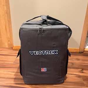 Könnte beinhalten: Graue Stofftasche mit schwarzem Reißverschluss und schwarzem Trageriemen. Die Tasche hat das Wort "VECTREX" in Weiß auf der Vorderseite gestickt und die Buchstaben "MB" in Rot und Blau.