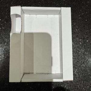 May include: A white cardboard box, rectangular in shape, with a cutout on one side. The box has folded sides and is designed for packaging or storage. The box is empty and sits on a dark surface.