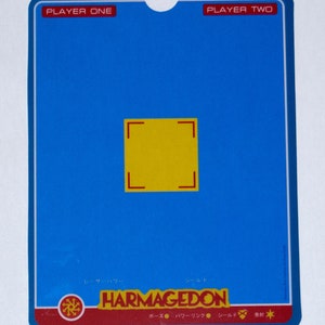 Peut inclure: Une carte bleue avec les mentions "PLAYER ONE" et "PLAYER TWO" en haut. La carte présente un carré jaune au centre avec des crochets d'angle rouges. Le bas de la carte affiche le mot "HARMAGEDON" en rouge et du texte japonais.