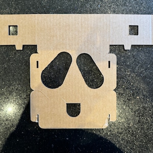 Może przedstawiać: Tekturowy wykrój twarzy z dwoma dużymi otworami na oczy i mniejszym otworem na usta. Wykrój jest brązowy i ma szorstką fakturę.