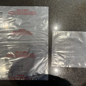 May include: Two transparent plastic bags, one rectangular and one square, are shown on a dark surface. The larger bag has red text, including the word "CAUTION". The bags are clear, revealing the surface below.