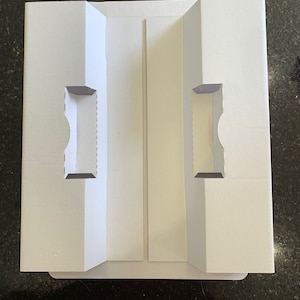 Puede incluir: Caja de cartón blanca con dos ranuras recortadas para guardar artículos. La caja está doblada por la mitad y tiene un pliegue en el medio.
