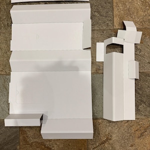 May include: White cardboard packaging components, including boxes and flaps, are arranged on a brown and gray tiled surface. The components are unfolded and ready for assembly, suggesting a product packaging theme.